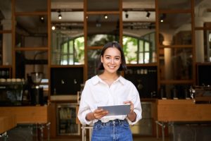 Portrait of asian woman, manager standing with tablet in front of cafe entrance, welcomes guests.